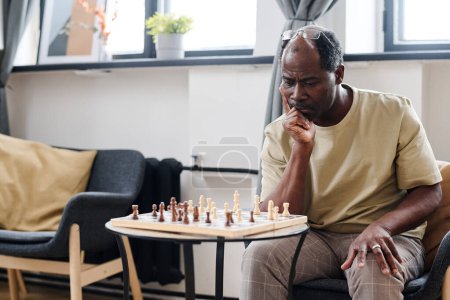 Téléchargez les photos : Pensive retired man of African ethnicity looking at chess board on small round table while thinking of next move - en image libre de droit