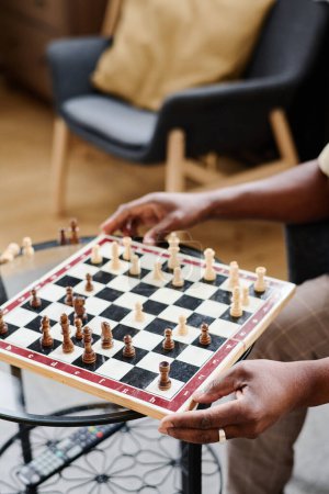 Foto de Hands of senior African American man holding chess board with black and white wooden figures while sitting by small glass table - Imagen libre de derechos