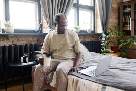 Photo for Serious retired black man with cup of coffee looking through fresh online news reel on laptop screen while sitting on double bed in the morning - Royalty Free Image