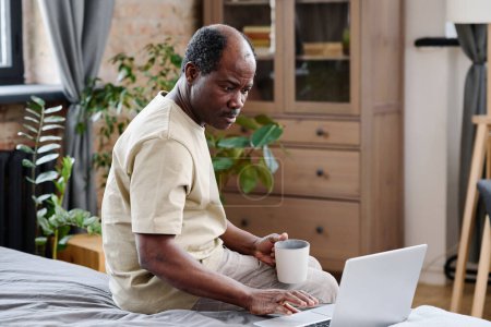 Téléchargez les photos : Pensionné afro-américain sérieux regardant l'écran d'ordinateur portable pendant la communication en ligne avec sa famille alors qu'il était assis sur le lit - en image libre de droit