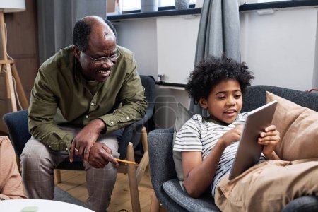 Photo for Happy senior African American man discussing online data with his cute grandson pointing at tablet screen while sitting in armchair - Royalty Free Image