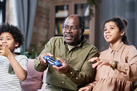 Téléchargez les photos : Des écoliers interculturels contemporains et leur grand-père afro-américain avec des joysticks jouant à un jeu de loisirs devant un téléviseur - en image libre de droit