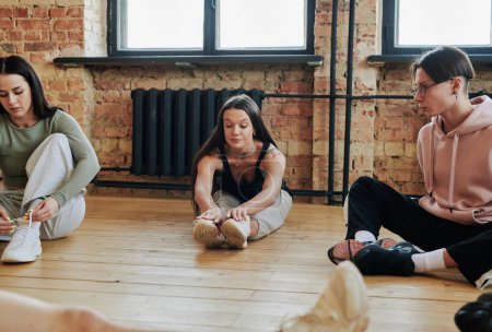 Foto de Active adolescent girls and guy sitting on the floor of spacious modern dance hall or studio and exercising before choreographic repetition - Imagen libre de derechos