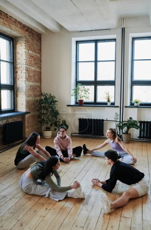 Téléchargez les photos : Groupe d'adolescents actifs assis sur le sol en bois d'une salle de danse ou d'un studio spacieux et pratiquant des exercices d'étirement et d'équilibre - en image libre de droit