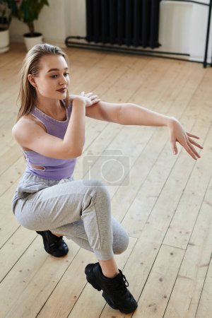 Téléchargez les photos : Happy female teenager in lilac tanktop and grey sweatpants performing modern dance while squatting on wooden floor in studio - en image libre de droit