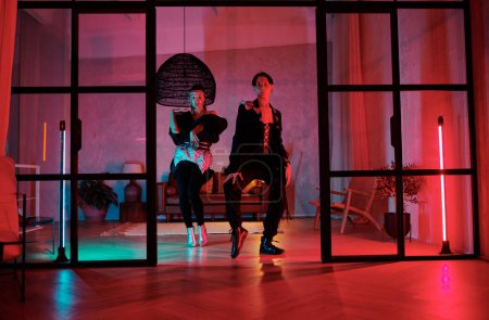 Téléchargez les photos : Two youthful dynamic dancers performing movements of vogue dance in studio or loft apartment lit by pink neon lamps in front of the door - en image libre de droit