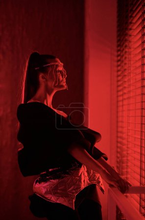 Photo for Side view of youthful girl with glamorous decoration or accessory on her face looking through venetian blinds during performance - Royalty Free Image