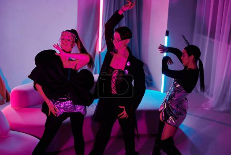 Téléchargez les photos : Groupe d'adolescentes contemporaines chic et mec appréciant la fête de balle de vogue à la maison et dansant dans le salon éclairé par des lampes au néon - en image libre de droit