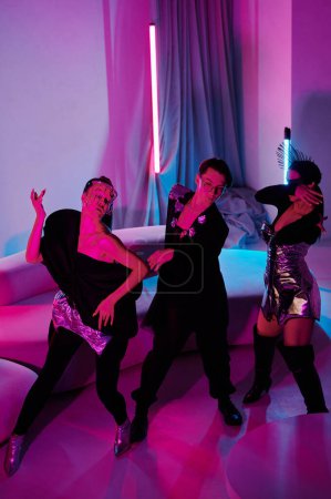 Téléchargez les photos : Group of dynamic teenage girls and guy in posh apparel dancing at home party against soft couch in living room lit by neon lamps - en image libre de droit
