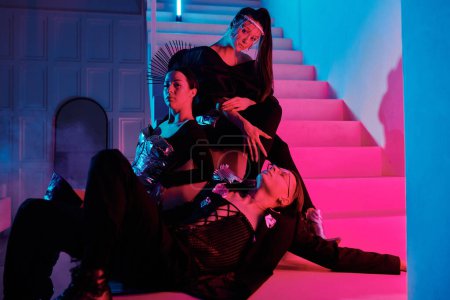 Téléchargez les photos : Posh adolescent gars et deux filles glamour dans des vêtements élégants détente sur l'escalier dans le studio moderne éclairé par la lumière rose et bleu néon - en image libre de droit