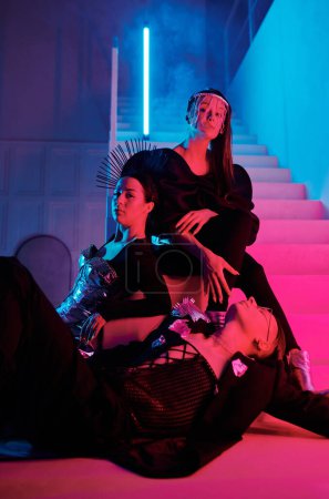 Téléchargez les photos : Group of three youthful vogue dance performers in posh attire posing on staircase of modern loft studio lit with pink and blue neon lights - en image libre de droit