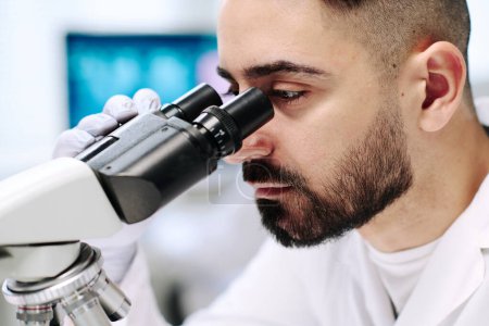 Foto de Part of face of young male researcher with microscope studying new virus while sitting by workplace against computer screen in laboratory - Imagen libre de derechos