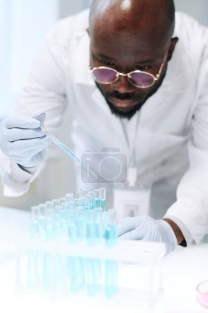 Téléchargez les photos : African American chemist mixing two liquid substances in chemical laboratory while dropping one of them into several flasks - en image libre de droit