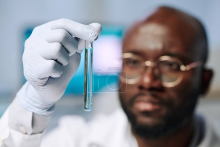 Photo for Gloved hand of African American male researcher holding flask with light blue liquid and looking at it during scientific investigation - Royalty Free Image