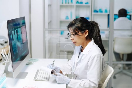 Photo for Contemporary young female researcher in whitecoat and gloves making scientific notes in notepad while sitting in front of computer - Royalty Free Image