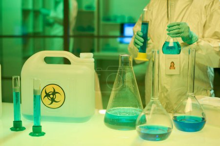 Foto de Contemporary clinician in coveralls and gloves holding two test tubes with blue liquid toxic substance while standing by workplace in lab - Imagen libre de derechos
