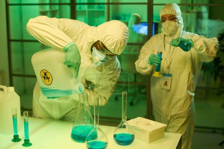 Hombre negro con batas protectoras mezclando sustancias líquidas en un tubo de ensayo grande contra su colega femenina haciendo otro experimento
