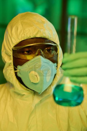 Photo for Young black man in protective coveralls looking at big test tube with blue liquid while studying characteristics of new chemical substance - Royalty Free Image