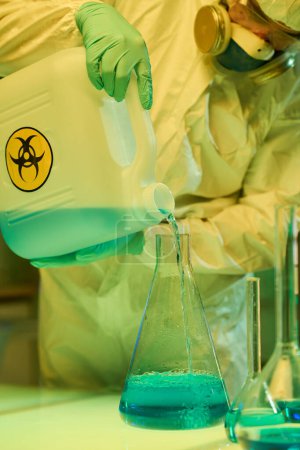 Foto de Primer plano del científico moderno en ropa de trabajo protectora mezclando sustancias líquidas mientras vierte una de ellas en un tubo de ensayo grande - Imagen libre de derechos