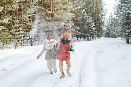 Foto de Cheerful African American family in winterwear having fun on snowy day while spending time in park or forest among evergreen trees - Imagen libre de derechos