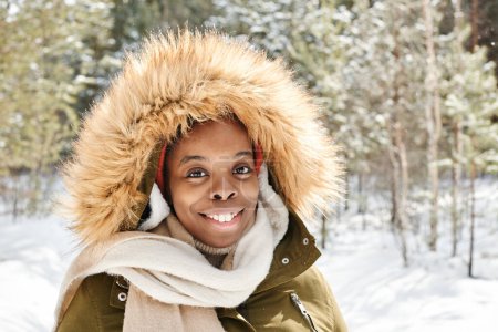 Foto de Happy young African American female in winter jacket and white woolen scarf looking at camera while spending time in park or forest - Imagen libre de derechos