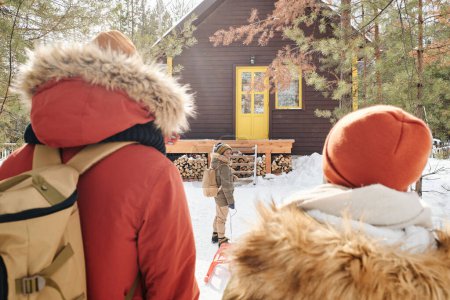 Foto de Rear view of young couple with backpacks and their little son with sledge moving towards their country house standing among pinetrees - Imagen libre de derechos