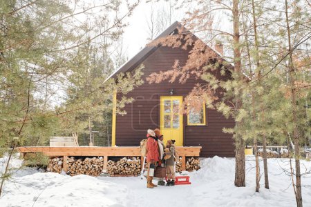 Téléchargez les photos : Famille contemporaine de parents et petit fils en vêtements d'hiver debout à côté de leur maison de campagne située dans les bois profonds parmi les pins - en image libre de droit