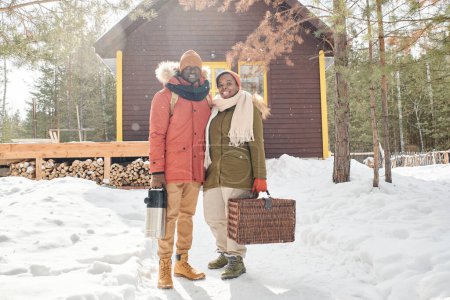 Young contemporary African American husband and wife in winterwear holding suitcase with household stuff and thermos for hot tea