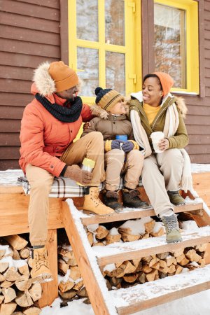 Photo for Happy African American family of three talking and having hot tea while sitting on wooden porch by their country house on winter day - Royalty Free Image