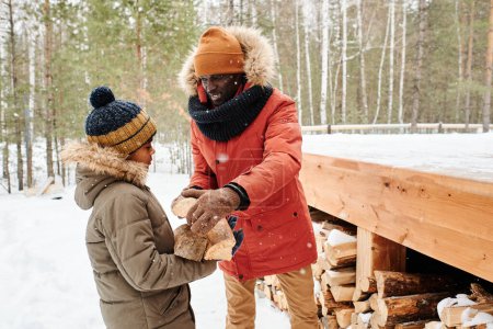 Foto de Young man in winterwear putting firewood on hands of his little son helping him bring wooden logs to their country house - Imagen libre de derechos