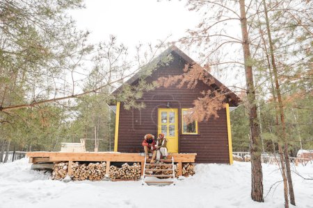 Téléchargez les photos : Maison de campagne en bois avec jeune couple affectueux assis sur son porche, ayant du thé chaud et parlant tout en se regardant - en image libre de droit