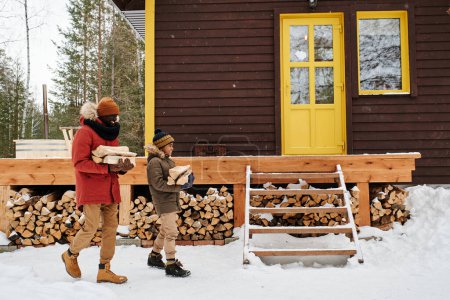 Téléchargez les photos : Heureux jeune homme noir et petit garçon en vêtements d'hiver décontractés chauds portant du bois de chauffage à leur maison de campagne située dans la forêt - en image libre de droit