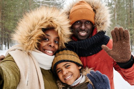 Téléchargez les photos : Famille afro-américaine de trois en vêtements d'hiver vous saluant tout en regardant la caméra dans le parc ou la forêt sur une journée d'hiver givrée à loisir - en image libre de droit