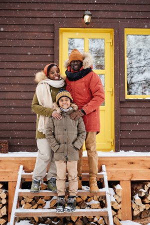 Téléchargez les photos : Heureux jeune couple et leur mignon fils en vêtements d'hiver debout sur un escalier en bois par le porche de leur maison de campagne - en image libre de droit
