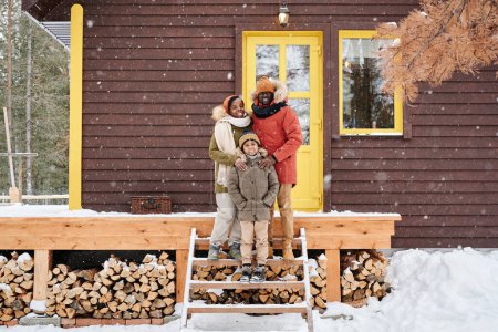 Foto de Tres miembros afroamericanos felices de la familia de pie junto al porche de la nueva casa de campo mientras disfruta del día de invierno nevado - Imagen libre de derechos