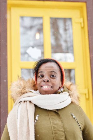 Foto de Joven viajera bastante femenina en ropa de invierno caliente de pie contra la puerta amarilla de la casa de campo o casa de campo y mirando a la cámara - Imagen libre de derechos