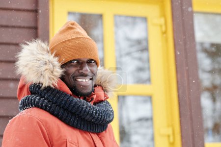 Foto de Young smiling black man in winter jacket, beanie hat and warm woolen scarf standing in front of camera against new country house - Imagen libre de derechos
