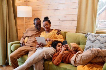 Foto de Feliz joven pareja cariñosa viendo vídeo en línea en la tableta mientras se relaja en el sofá verde suave junto a su hijo con el teléfono inteligente - Imagen libre de derechos