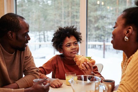 Téléchargez les photos : Mignon afro-américain garçon avec fait maison hotfog écoute sa mère par petit déjeuner tandis que la famille de trois assis à table contre grande fenêtre - en image libre de droit