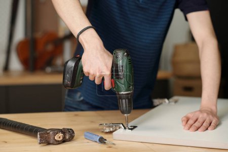 Foto de Hands of youthful boy holding electric drill while fixing hinge on door of kitchen cabinet while standing by workbench in garage - Imagen libre de derechos