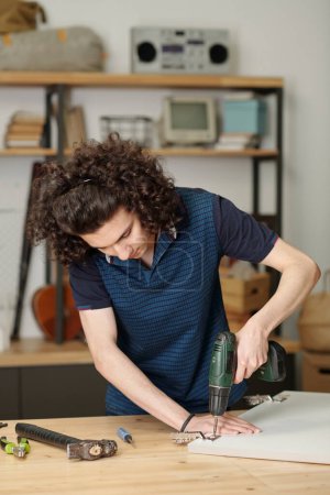 Photo for Young serious guy in blue shirt bending over table while drilling metallic hinge of plywood door of kitchen cabinet in garage of his house - Royalty Free Image