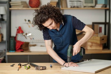 Foto de Accurate guy with dark wavy hair using electric drill for fixing metallic hinge on plywood door of kitchen cabinet while standing by table - Imagen libre de derechos