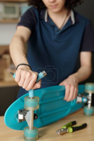 Foto de Manos de adolescente con llave inglesa o destornillador fijando ruedas de monopatín azul o reparándolo mientras trabaja por mesa o banco de trabajo - Imagen libre de derechos
