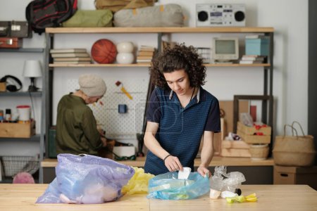 Téléchargez les photos : Teenage guy sorting waste and putting it into two large cellophane sacks while standing by table in garage against his grandmother - en image libre de droit