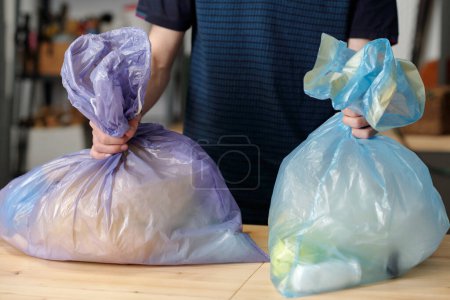 Photo for Two large cellophane sacks held by teenage guy standing by table in garage after sorting variety of waste into different bags - Royalty Free Image