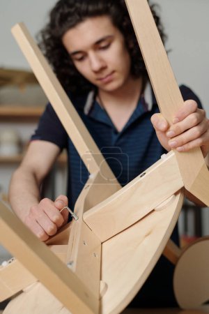 Foto de Teenage boy with handtool fixing back part of seat of wooden chair in garage while repairing it or finishing with new item for his family - Imagen libre de derechos