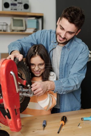 Foto de Young man showing his little daughter how to fix part of bicycle wheel while standing next to her by table with the vehicle in garage - Imagen libre de derechos