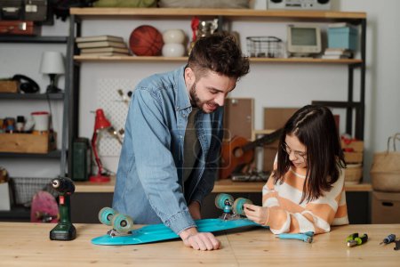 Téléchargez les photos : Jolie fille vérifier roues de skateboard bleu après les avoir fixés avec l'aide et les conseils de son père tout en étant debout à côté de la table en bois - en image libre de droit