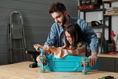 Téléchargez les photos : Cute smiling girl with handtool fixing wheels of skateboard on wooden table in garage while young man in denim shirt helping her - en image libre de droit