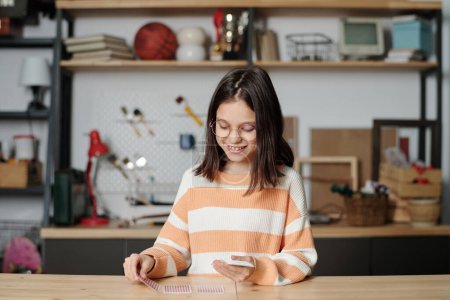 Foto de Cute smiling little girl in eyeglasses and striped pullover putting row of game cards on wooden table while playing at leisure in garage - Imagen libre de derechos